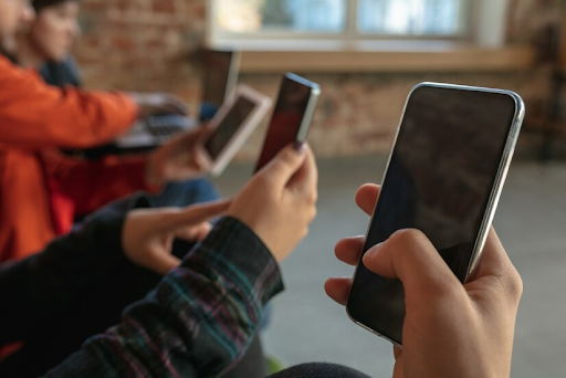 Imagem close-up nas mãos de três pessoas segurando smartphones. Ao fundo podemos ver outras duas usando um notebook.
