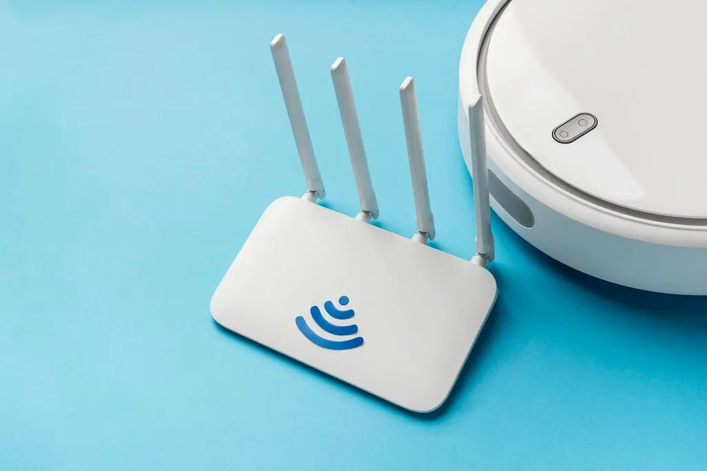 Roteador Wi-Fi 6: 5 Sinais para dar um Upgrade na Conexão! 2 Vista de ângulo alto de um roteador Wi-Fi branco deitado em uma superfície azul, com três antenas brancas levantadas. Um adesivo com um símbolo de Wi-Fi azul em forma de onda está no centro.